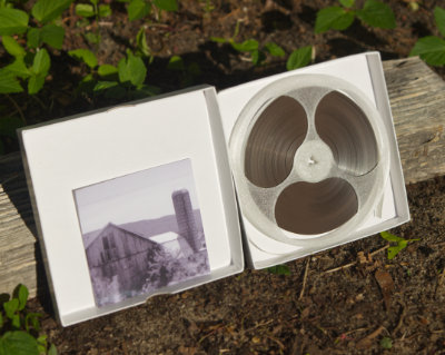 The insides of a five inch tape reel box containing an icy translucent reel of tape and a small black and white photograph of a barn and silo, rested against a log of firewood in front of some plants